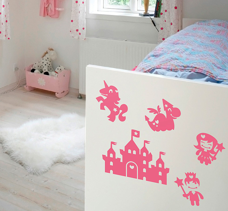 Photo of the end of a bed in a nursery. The bed is decorated with pink wallstickers drawn in silhouette. The wallstrickers depicts princesses, adventure castles, wizards and unicorns.