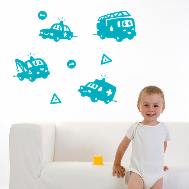 Baby in front of a sofa and a white wall with some turquoise wallstickers depicting cute rescue vehicles with eyes and a smiling mouth. Ther is a police car, a firetruck, an ambulance and a tow truck.