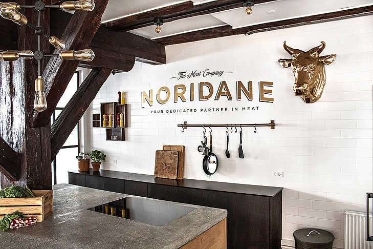 Interior decoration of show kitchen at a meat wholesaler in Copenhagen. In the foreground you can see a kitchen with a tabletop of concrete. Bulps are hanging from the ceiling and the kitche are built in raw oak wood. The wall in the background is covered in white metro tiles. On the wall there is a company logo in gold and a big golden bulls head.