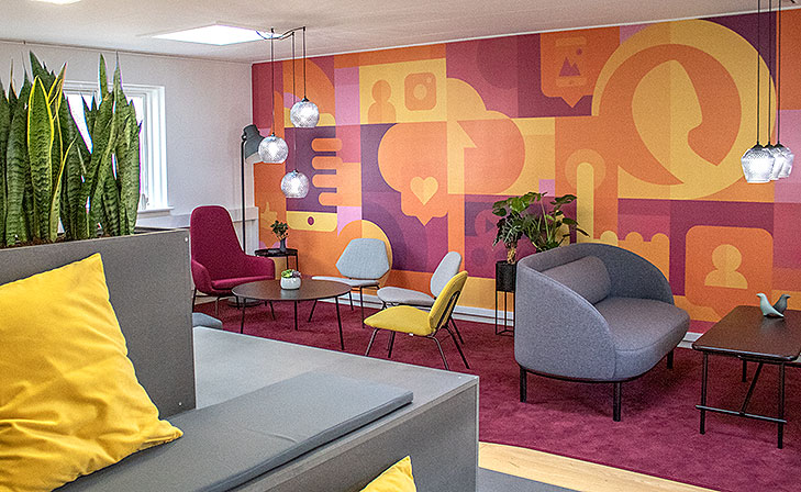 Lounge interior design seen from the right. In the foreground there is a customized lounge furniture with terraces, a yellow cushion and built in fower pots. Behind it there is a big purple carpet. To the right there is two Fuuga sofas with black legs and a Peter Boy sofa table. To the left there is a round black lounge table, yellow lounge chairs from Woud and purple armchairs from Normann Copenhagen. On the wall behind gthe lounge there is a big floor to ceiling wall painting in yellow, orange and purple colors. The wallpainting is abstract with recognisable elements like a cellphone, social media icons, communicatuon and app development. Both sofa tables have glas pendant lamps with crystal patterns hanging over them.