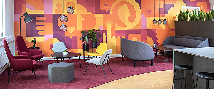 Lounge indretning set fra venstre side. I forgrunden ses et specialbygget terrasse bar bord i gråt MDF med sorte barstole i sort træ og med tynde sorte stålben. Bagved ses et stort bordeaux farvet gulvtæppe. Til højre er der et sofa arrangement med to grå Fuuga sofaer med sorte ben og et sort Peter Boy sofa bord. Til venstre er der et rund sort lounge bord med grå og gule lounge stole fra Woud og lilla lænestole fra Normann Copenhagen. På væggen bag loungen ses et gulv til loft vægmaleri med store farveflader i gule, orange og lilla nuancer. Vægmaleriet er abstrakt med enkelte genkendelige elementer som mobiltelefoner, ikoner fra sociale medier, telekommunikation og app udvikling. Over begge sofaborde hænger pendler i glas med krystal-slebet mønster. I baggrunden til højre ses en specialbygget høj blomsterkasse.