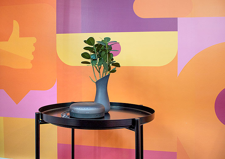 Nærbillede af et sort sidebord fra loungeindretning med lille potteplante og B&O Play højtaler. I baggrunden ses et gulv til loft vægmaleri i gule, orange og lilla nuancer.